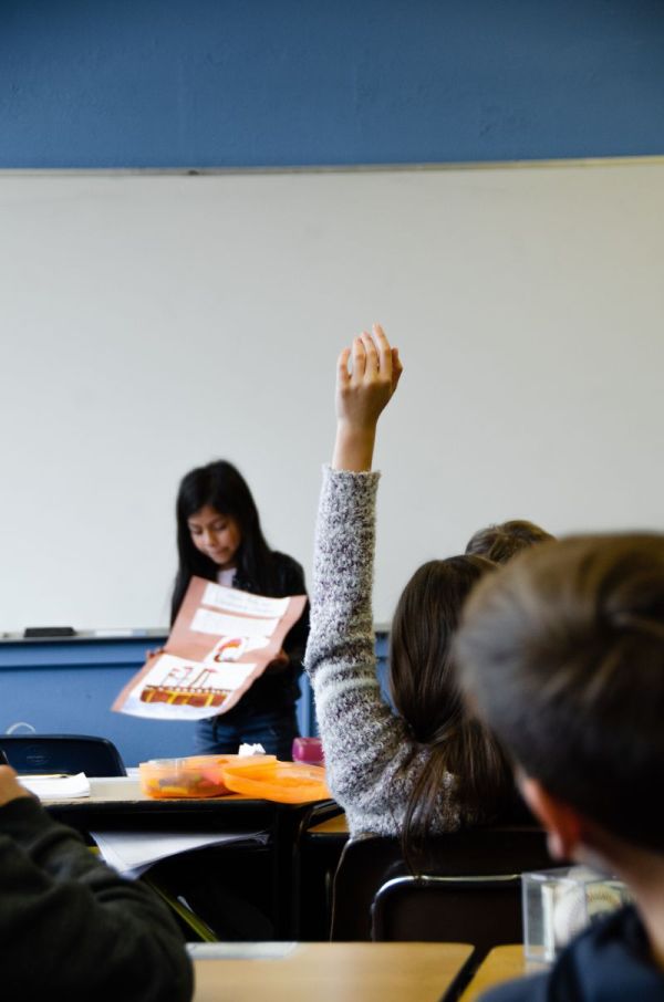 fetita bruneta care prezinta un proiect la cursul de engleza in sala de clasa in fata colegilor care stau in banci